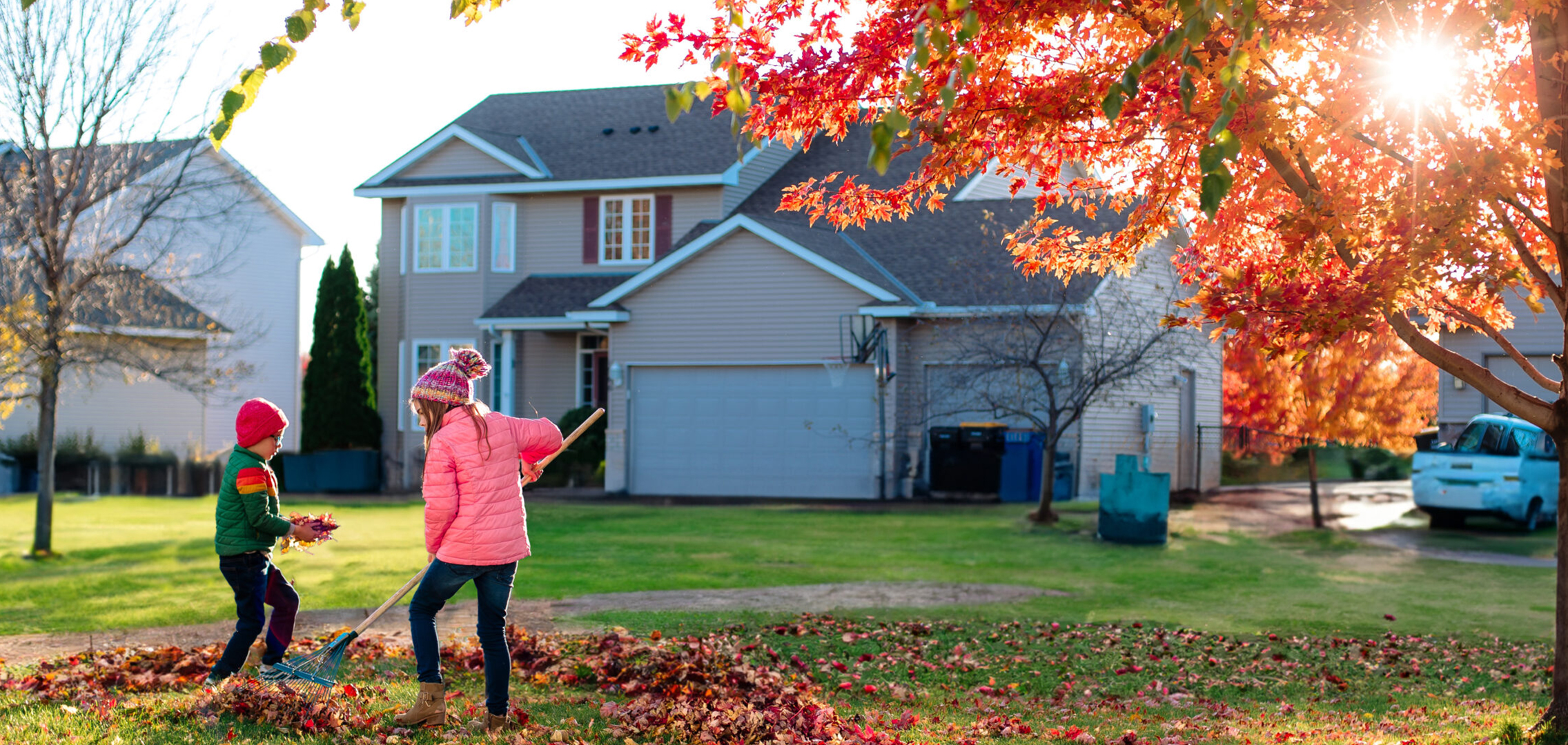 Residential house in autumn; maple tree with red leaves in front yard and two children raking leaves.